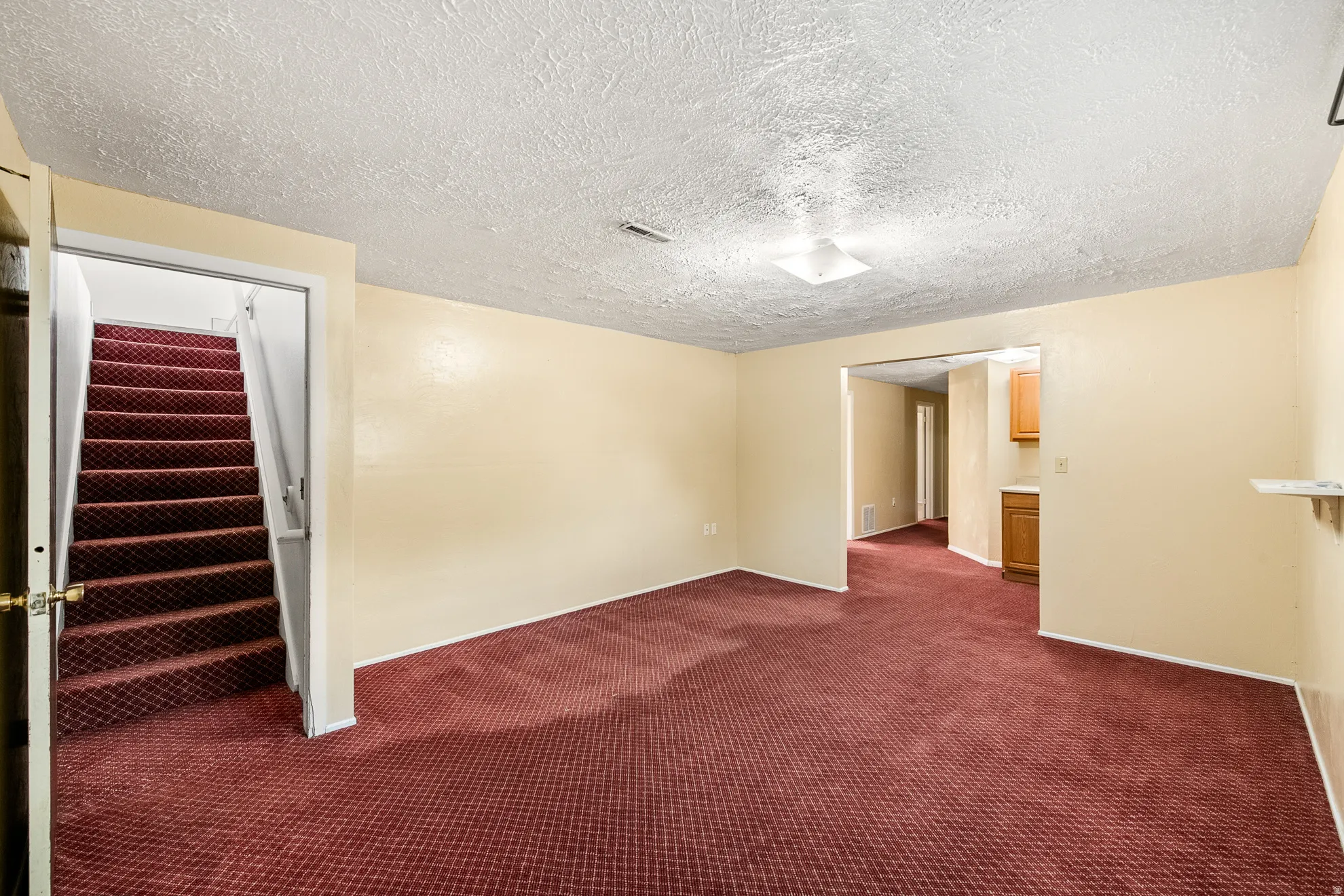 Carpeted room featuring a ceiling-mounted light fixture, light-colored walls, and a staircase with patterned carpeting