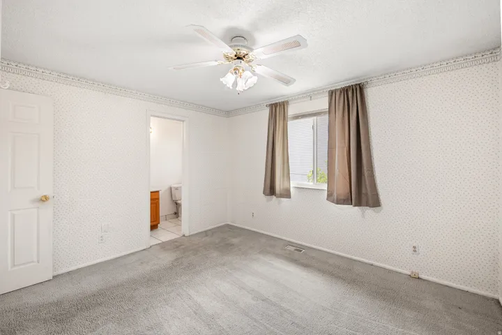 Carpeted room featuring a ceiling fan with light fixture, patterned wallpaper, a window with exterior visibility, and a doorway leading to a bathroom with a vanity