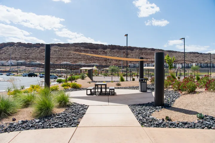 Community common area featuring a concrete pathway, a shaded picnic area with a dark-toned table and benches, and a large shade sail