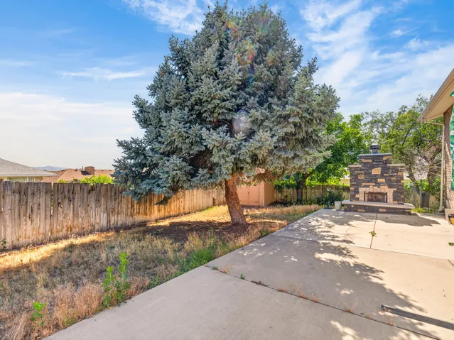 Spacious concrete patio with an outdoor stone-clad fireplace and a mature evergreen tree