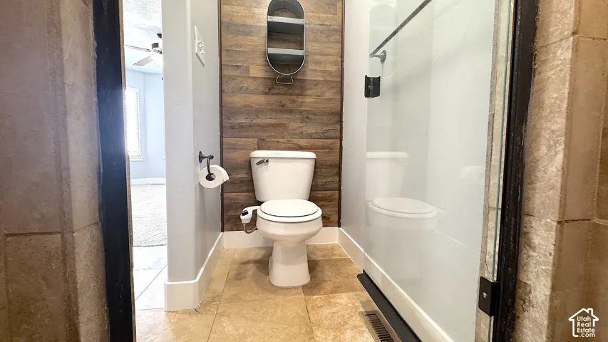Primary bathroom featuring a walk-in shower with clear glass enclosure, private toilet area, tile flooring, and a wood accent wall.