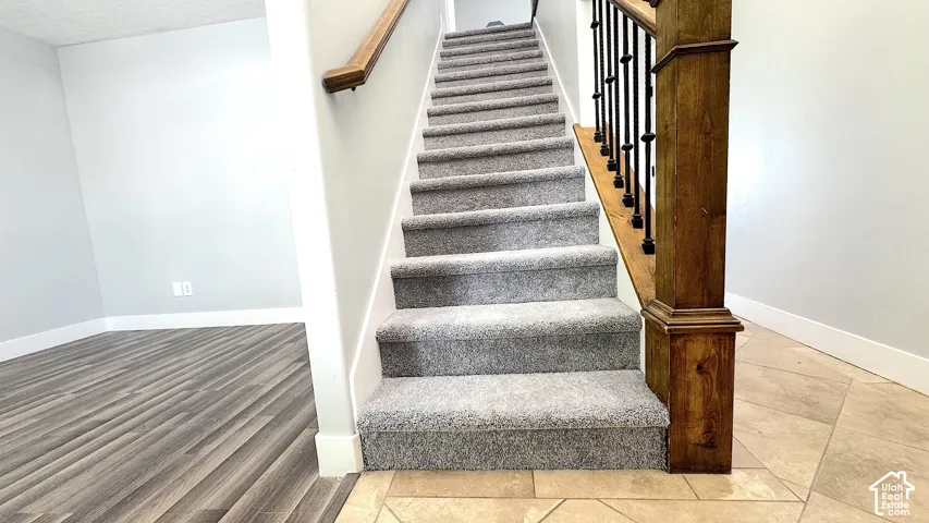 Carpeted staircase with a wood handrail and dark metal balusters, anchored by a substantial wood newel post. Office just off to the left.