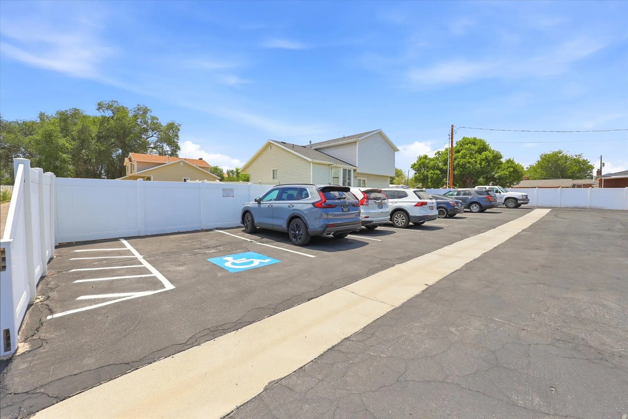 Paved parking area featuring multiple designated parking spaces, a white privacy fence, and a marked accessible parking space