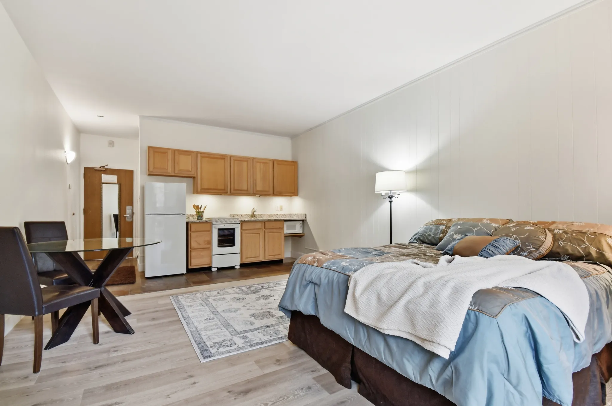 Studio living area featuring wood-finish flooring, a full kitchen with light wood cabinetry and appliances, and a dedicated dining space with a glass-top table