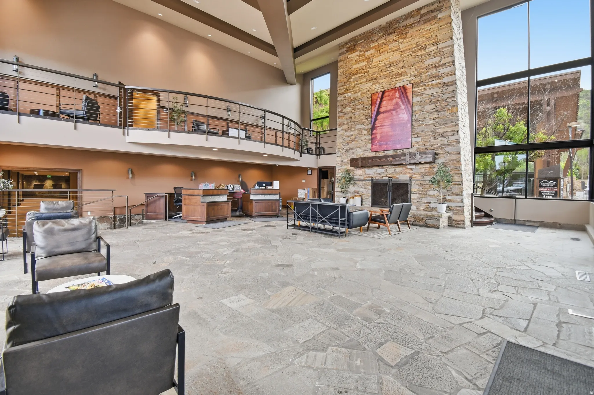 Spacious foyer with flagstone flooring, a two-story stone fireplace, expansive window walls, and a visible mezzanine with metal railings