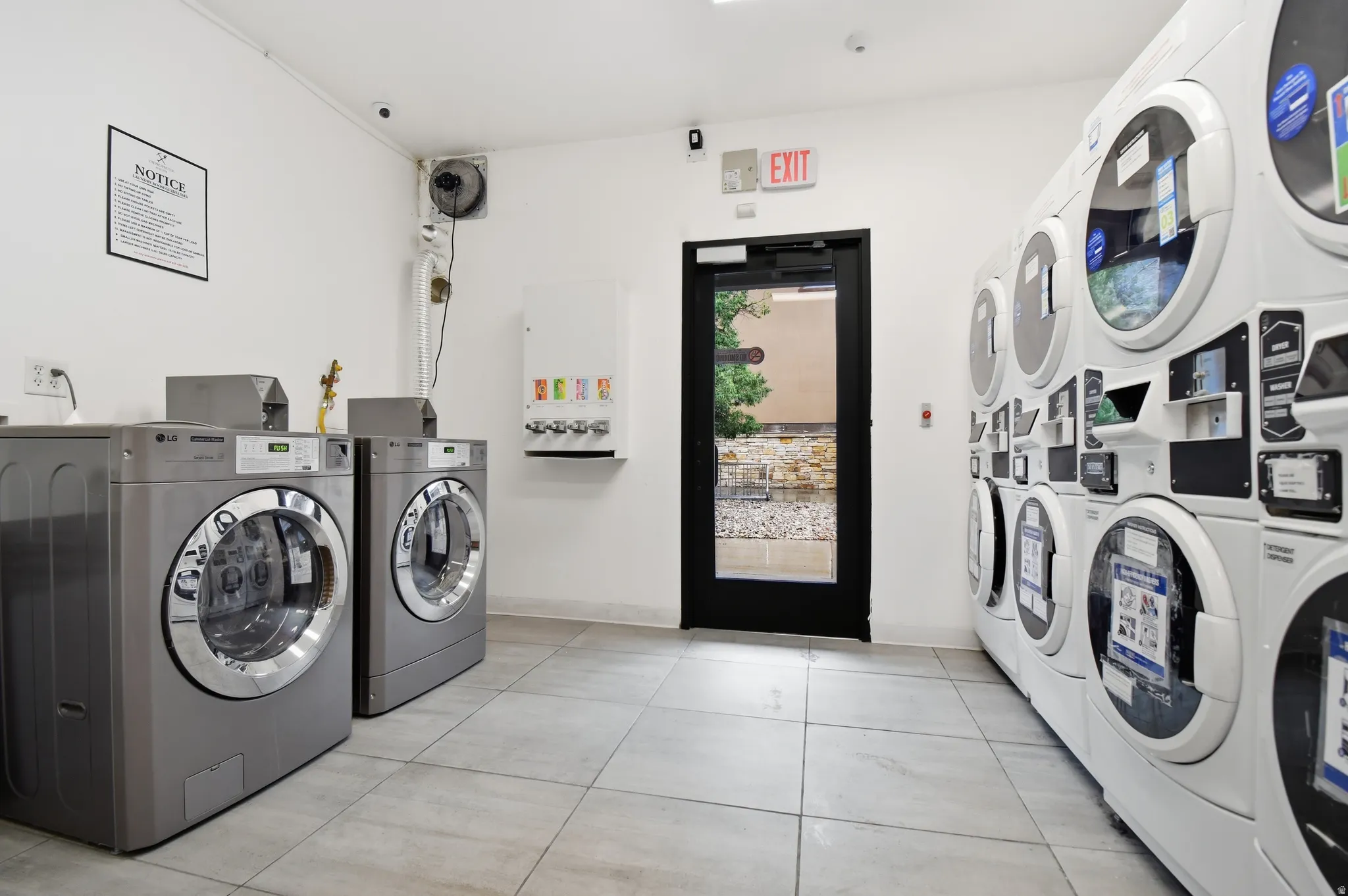 Dedicated laundry room featuring multiple front-loading washers and dryers, light-colored tile flooring, white walls, and a black-framed glass exit door