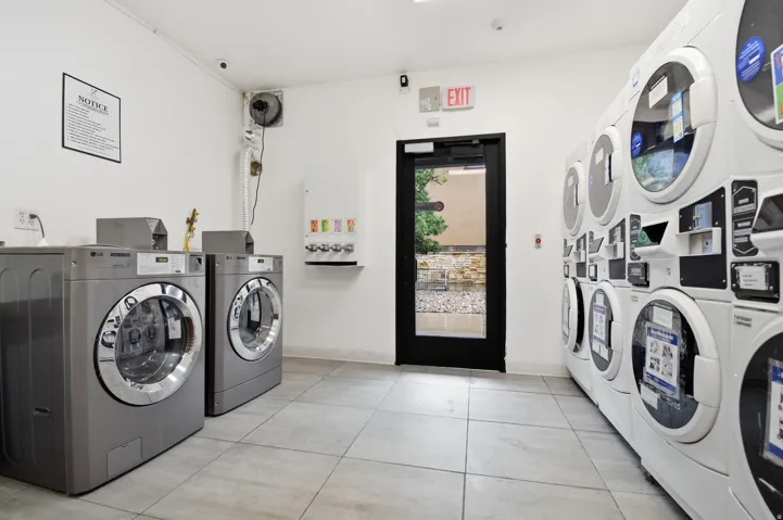 Dedicated laundry room featuring multiple front-loading washers and dryers, light-colored tile flooring, white walls, and a black-framed glass exit door