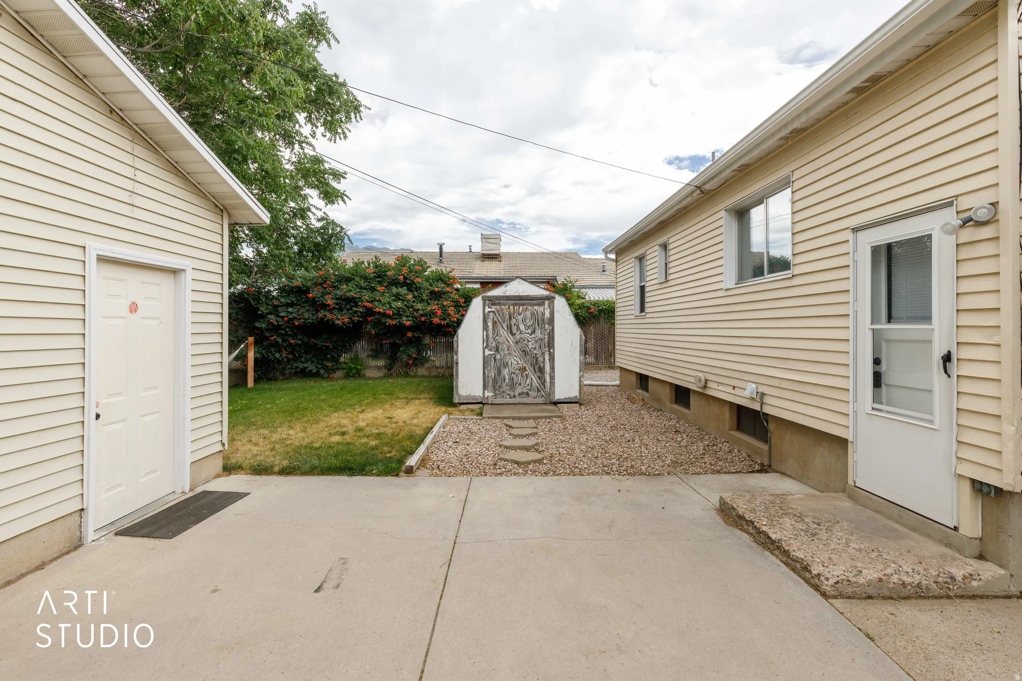 Concrete patio with gravel and a storage shed, bordered by a lawn area and mature trees