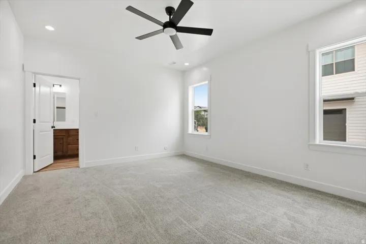Carpeted room featuring a ceiling fan with integrated lighting, two windows, and a doorway leading to a bathroom with a wood-finish vanity