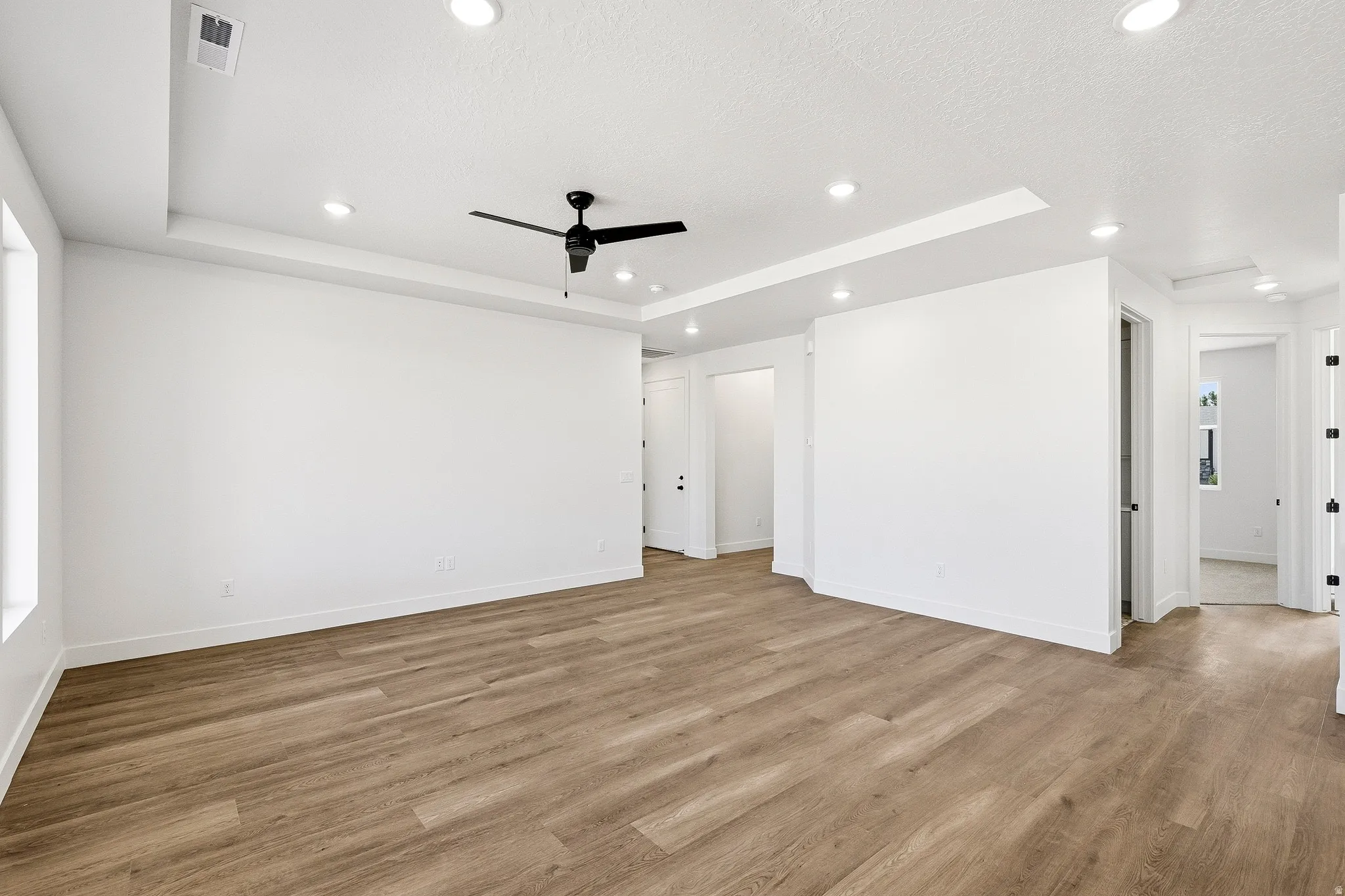 Spacious room featuring wood-finish flooring, white walls, and a recessed ceiling with a black ceiling fan and integrated lighting