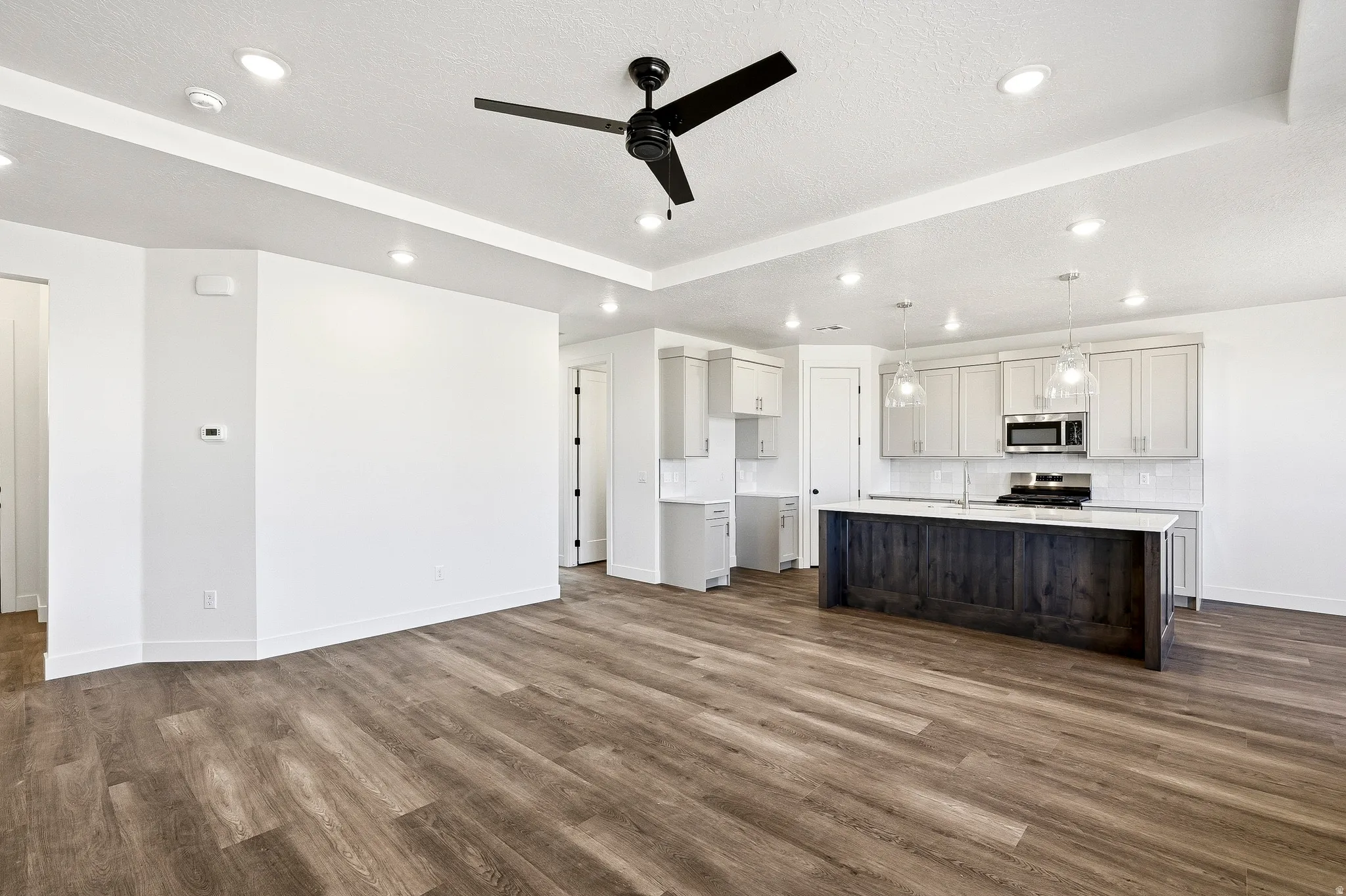 Open-concept living area featuring wood-finish flooring, a modern black ceiling fan, recessed lighting, a kitchen island with dark wood-finish paneling, light gray cabinetry, and stainless steel appliances