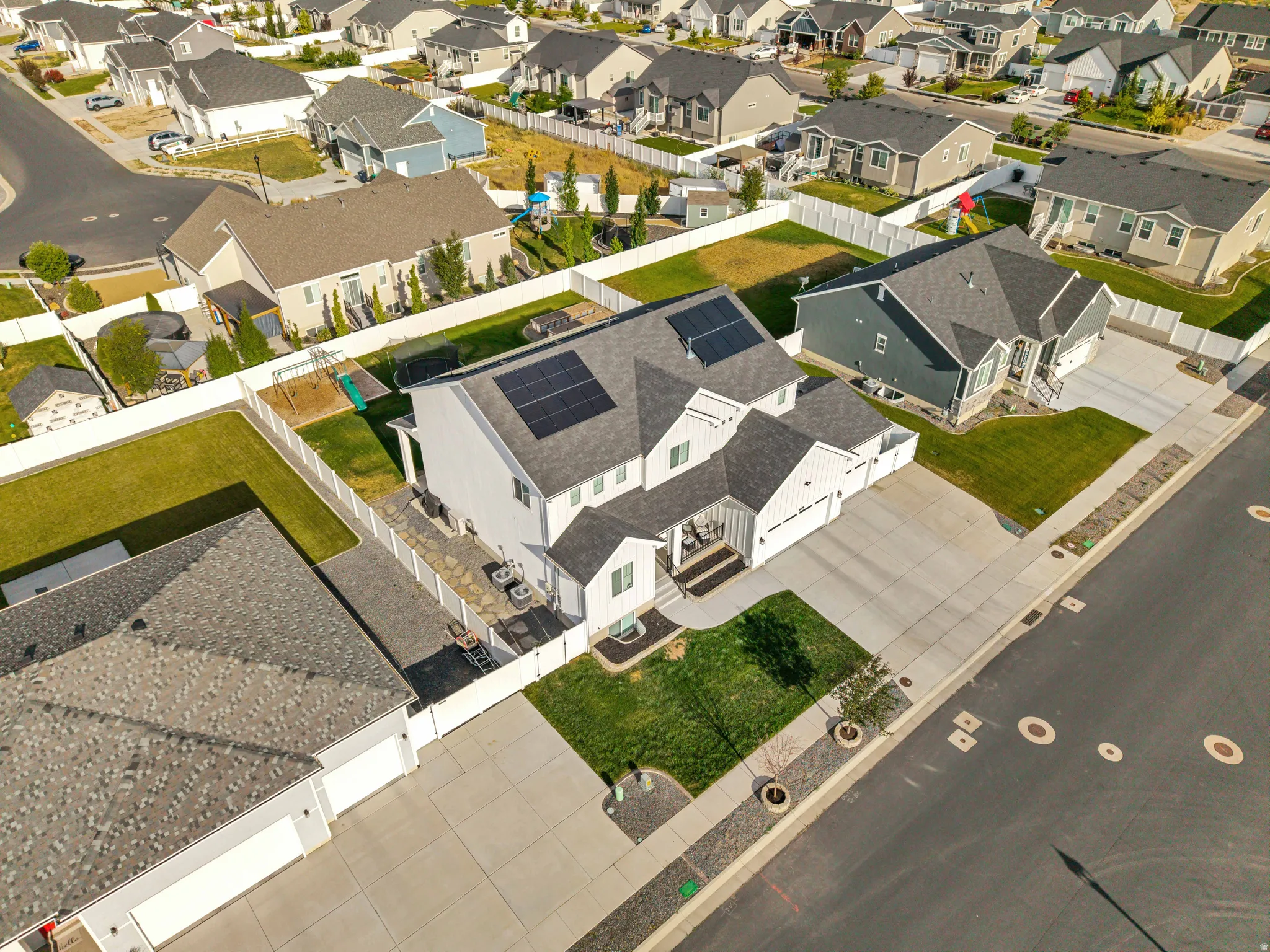 Contemporary white siding residence featuring a dark shingle roof with integrated solar panels, an attached garage, a spacious concrete driveway, and a fenced backyard with a patio