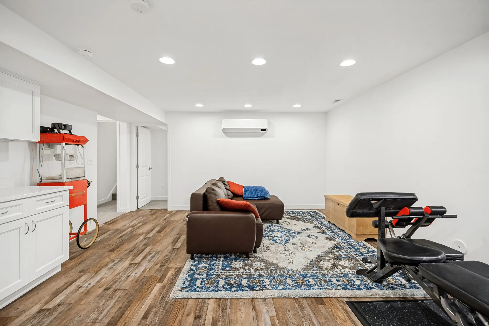 Open-concept recreation area featuring wood-finish flooring, white cabinetry with a light-toned countertop, recessed lighting, and a wall-mounted climate control unit