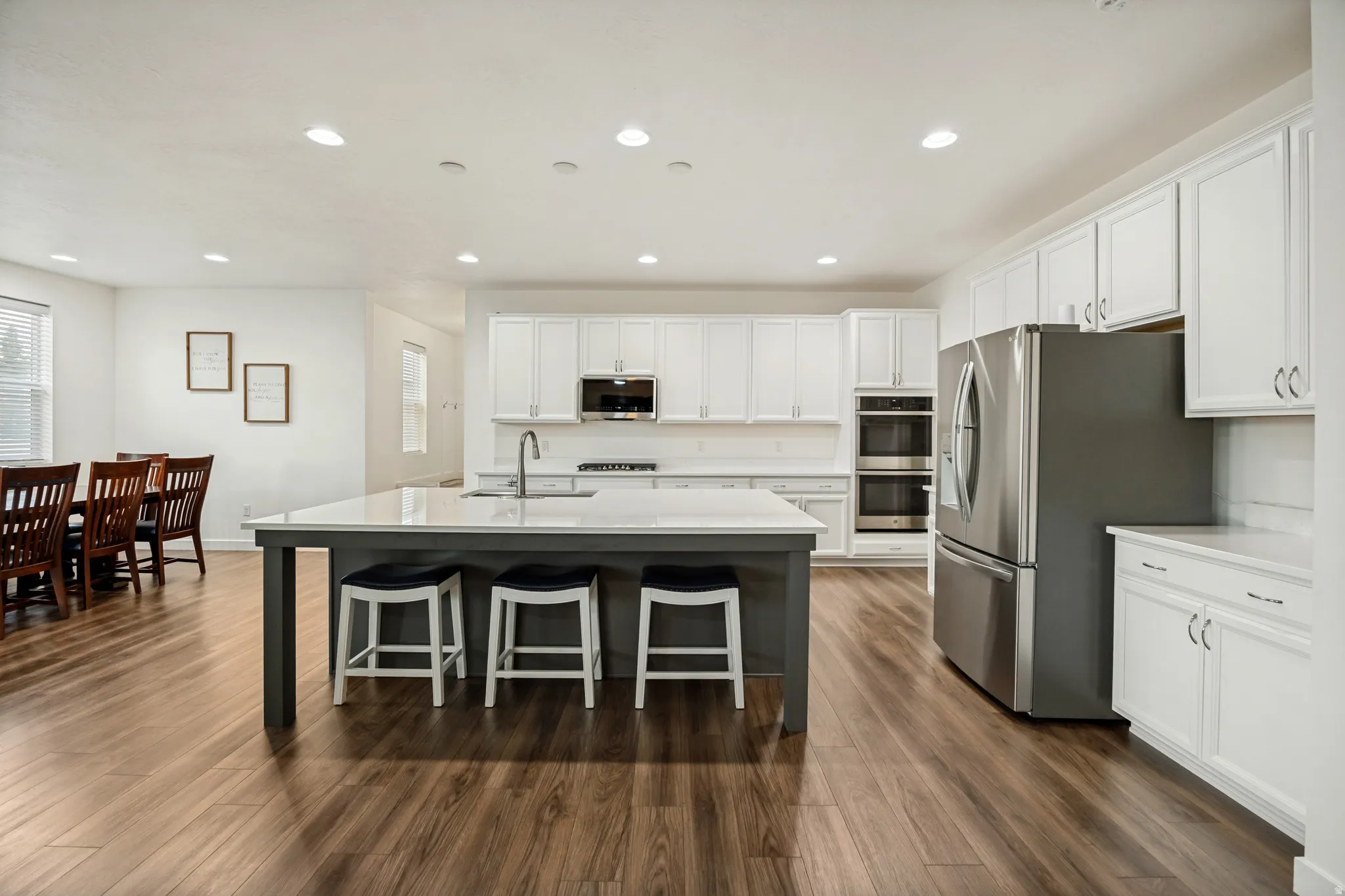 Open-concept kitchen featuring an island with seating, white shaker cabinetry, stainless steel appliances including a double oven and refrigerator, and wood-finish flooring