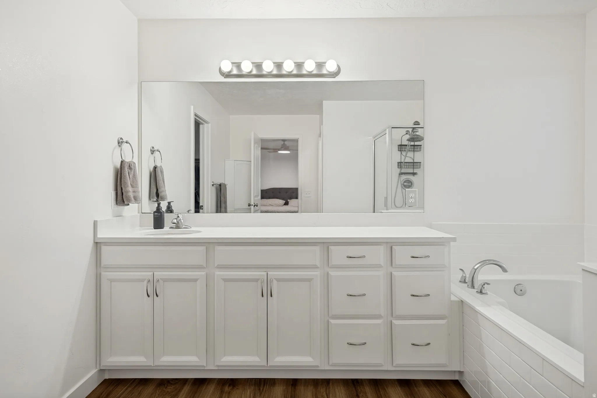 Bathroom featuring a double vanity with white cabinetry and chrome hardware, a large mirror, a chrome light fixture with five bulbs, a built-in bathtub with white tile surround, and wood-finish flooring