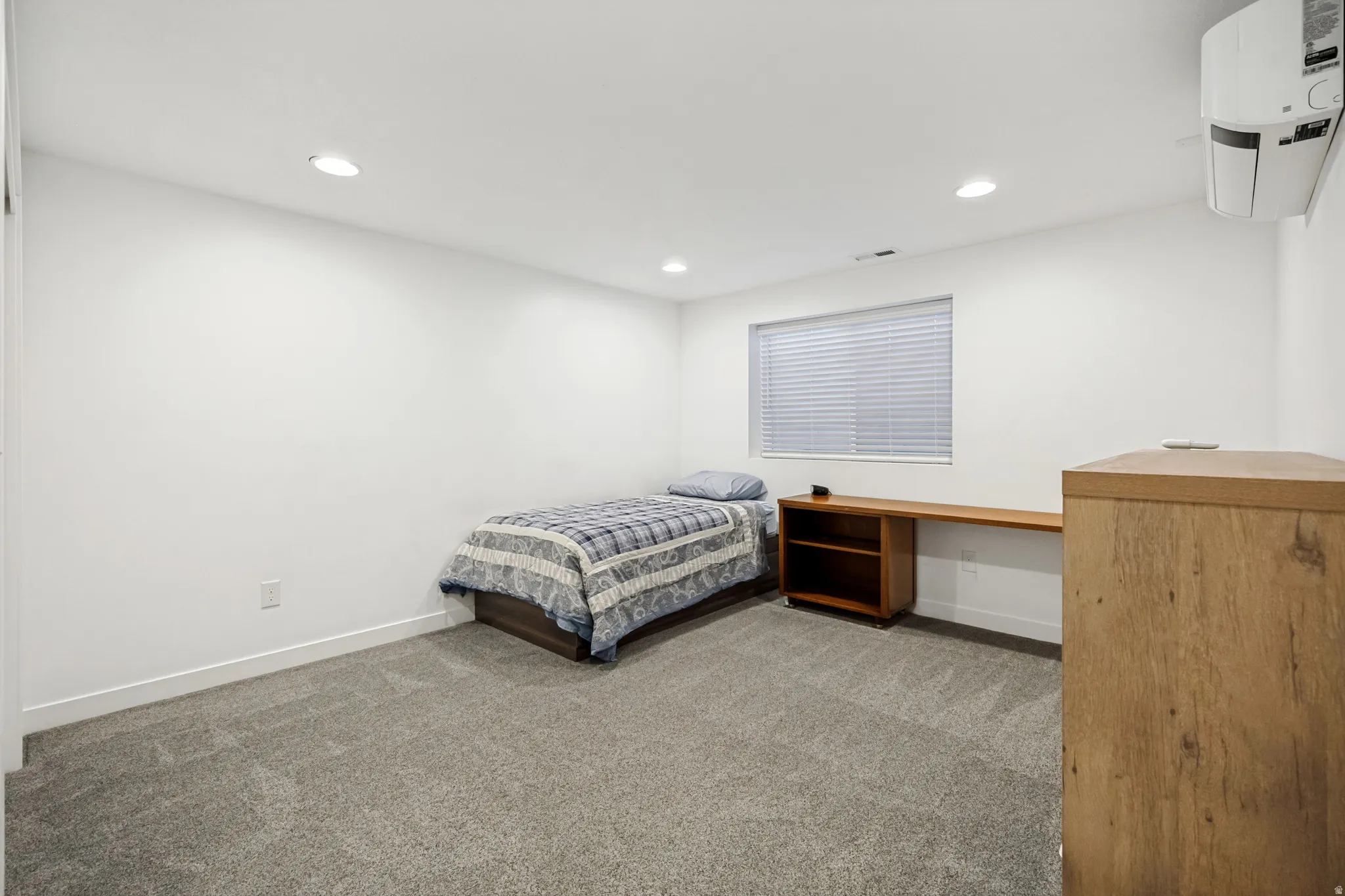 Carpeted room featuring recessed lighting, a built-in wood-finish desk with shelving, a window with horizontal blinds, and a wall-mounted mini-split unit