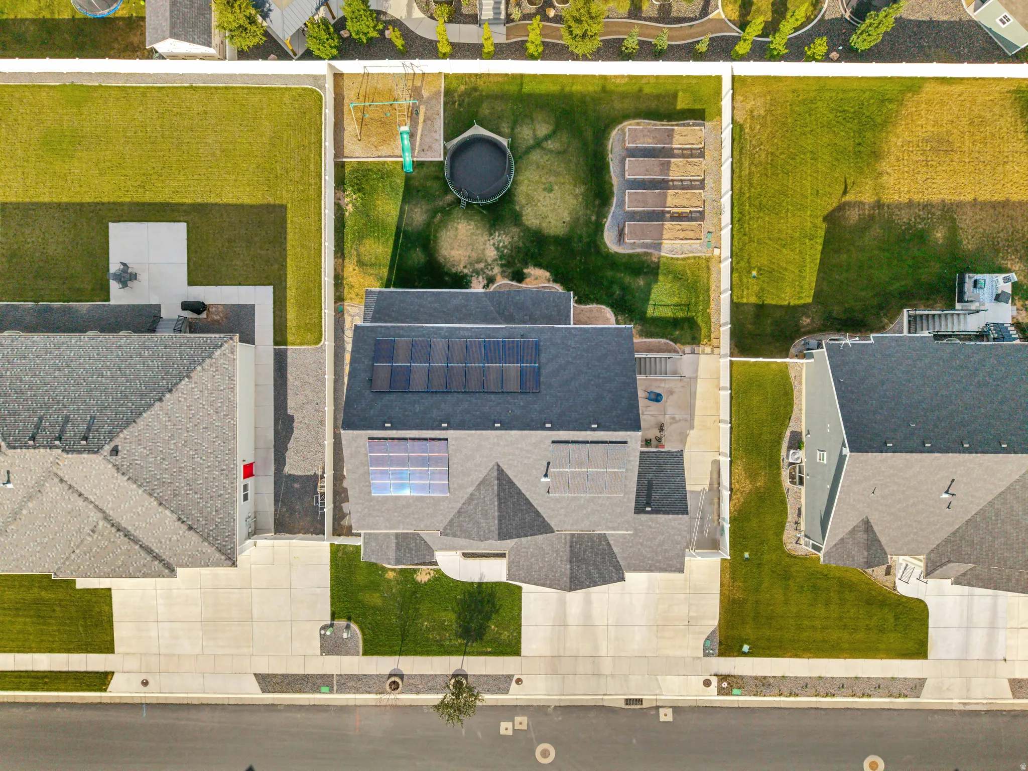 Aerial view of a residential property featuring solar panels on the roof, a fenced backyard with raised garden beds, and a concrete patio