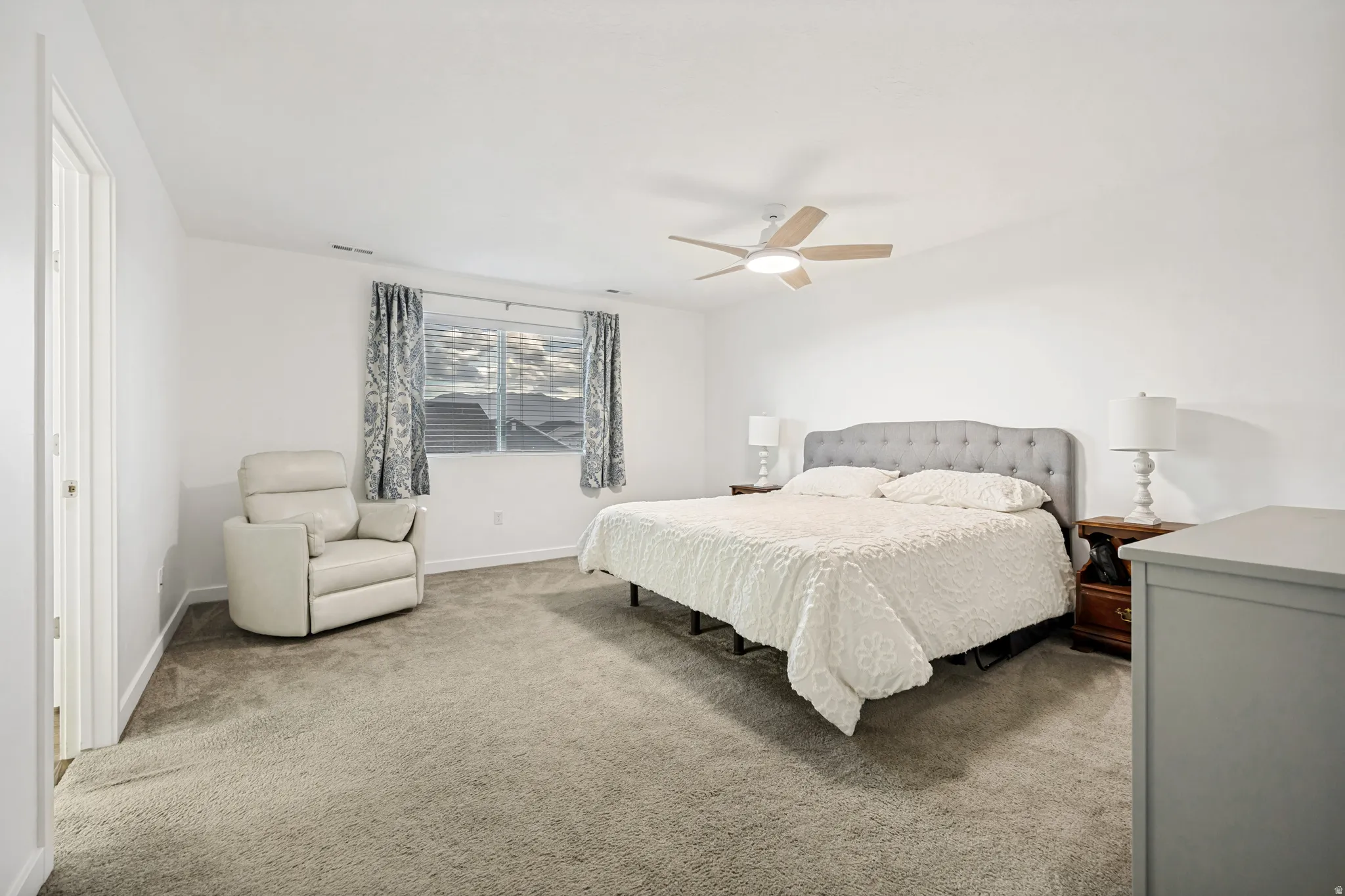 Carpeted bedroom with a ceiling fan light fixture, two windows with blinds and patterned curtains, and a light gray tufted headboard
