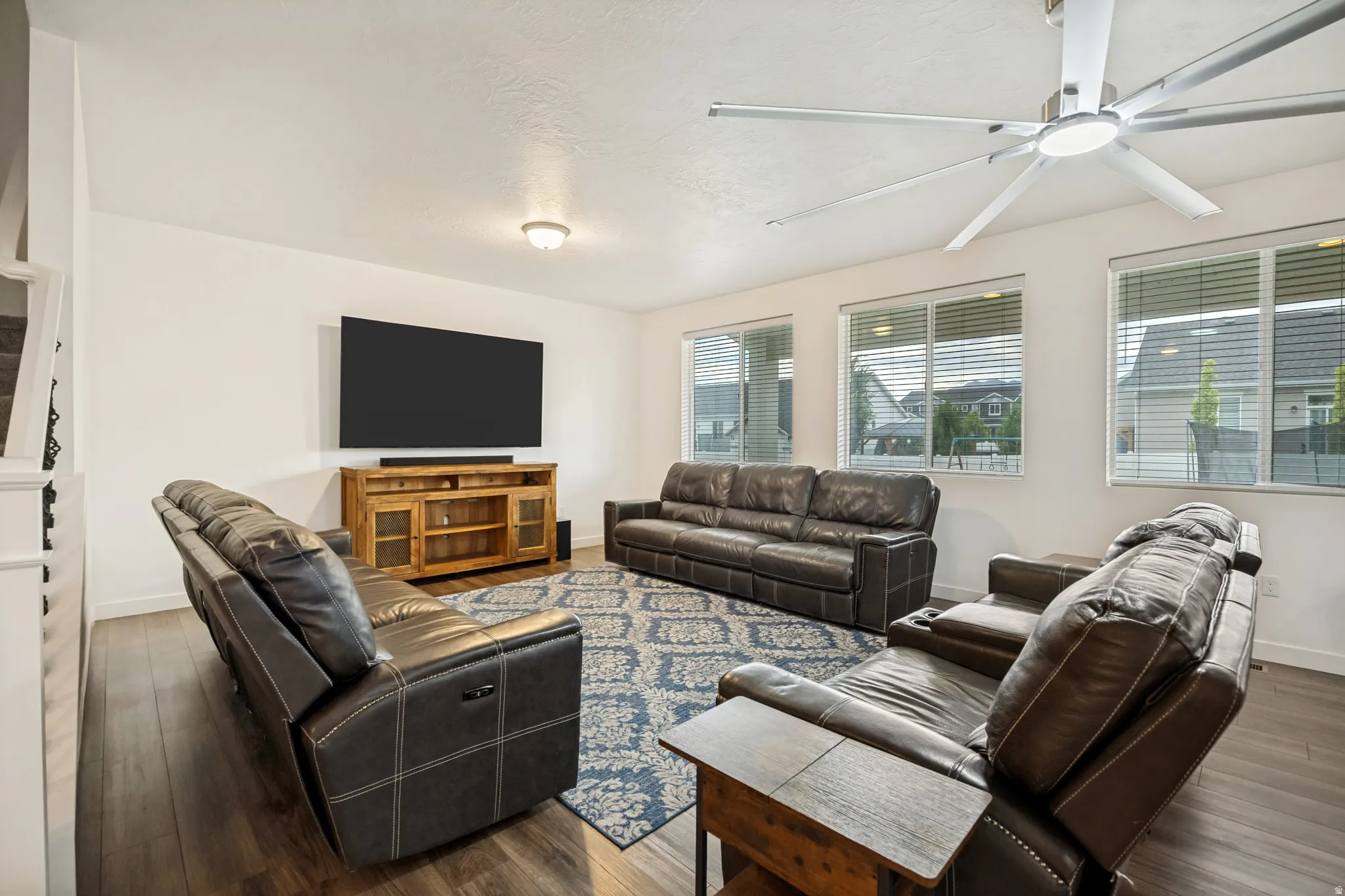 Spacious entertainment room featuring wood-finish flooring, a contemporary ceiling fan, and multiple windows with blinds