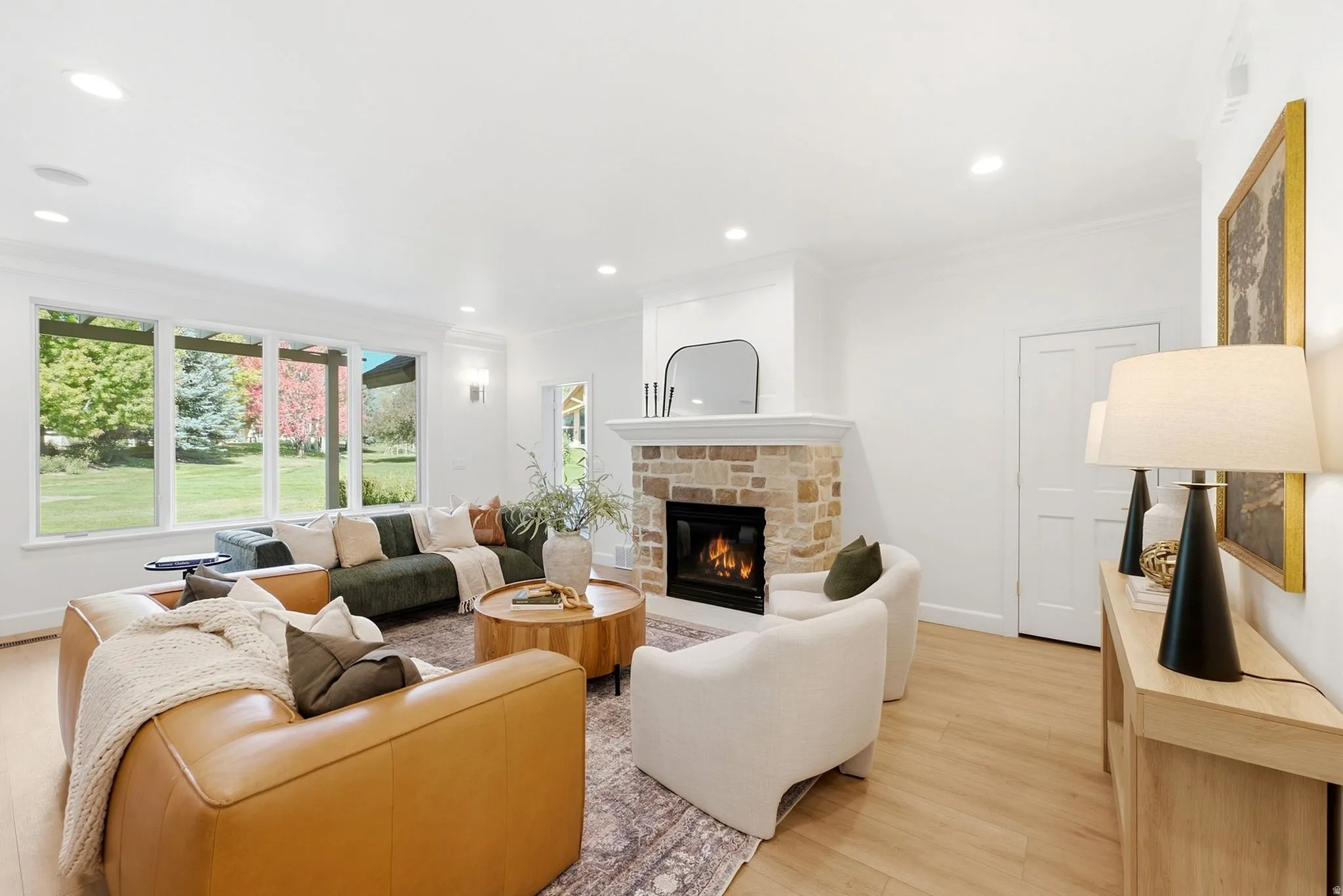 Light wood-finish flooring, stone-clad fireplace, large windows, recessed ceiling lighting, and crown molding