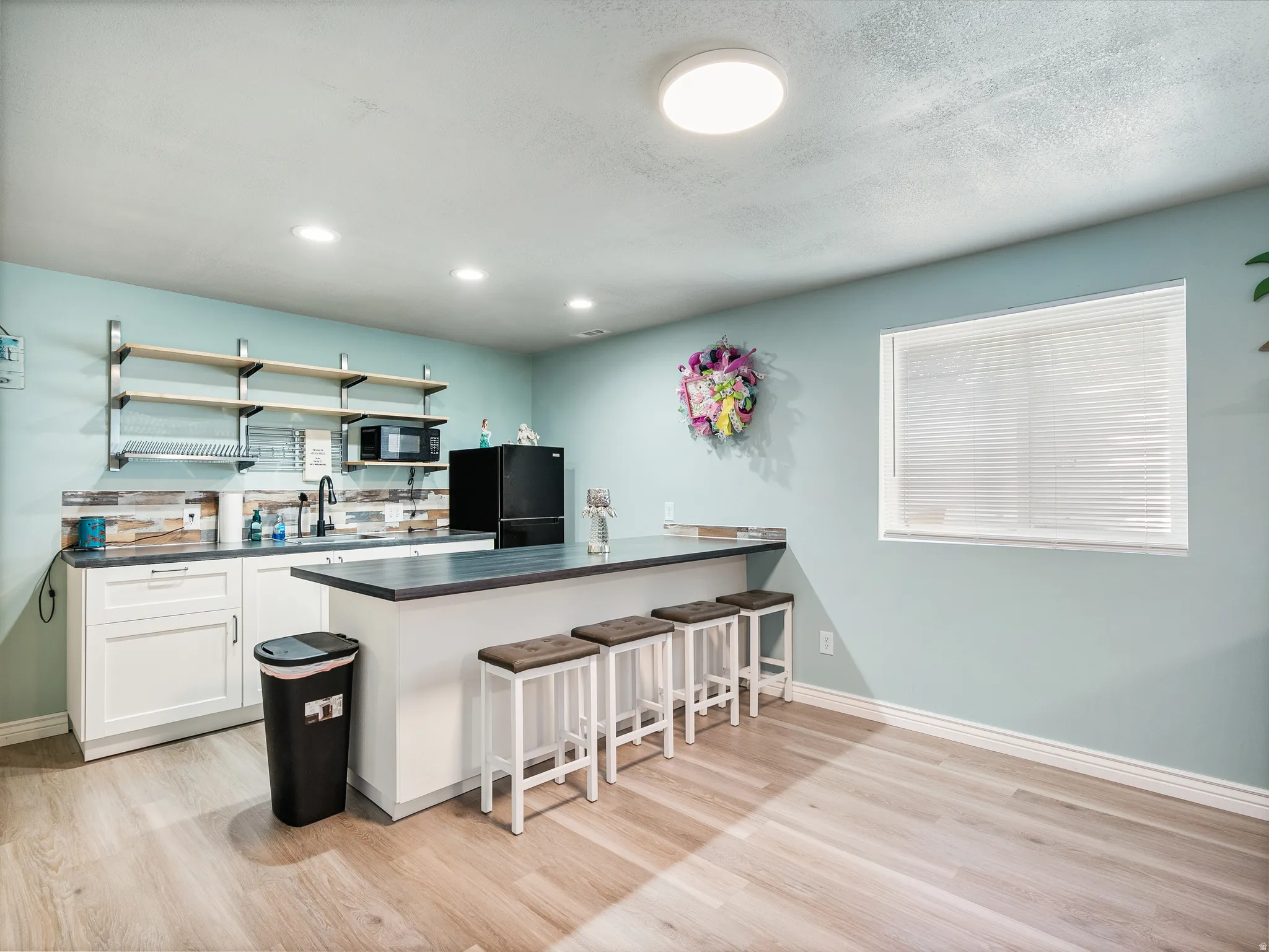 Kitchenette featuring light blue walls, wood-finish flooring, white cabinetry, open shelving, and a built-in countertop bar with seating