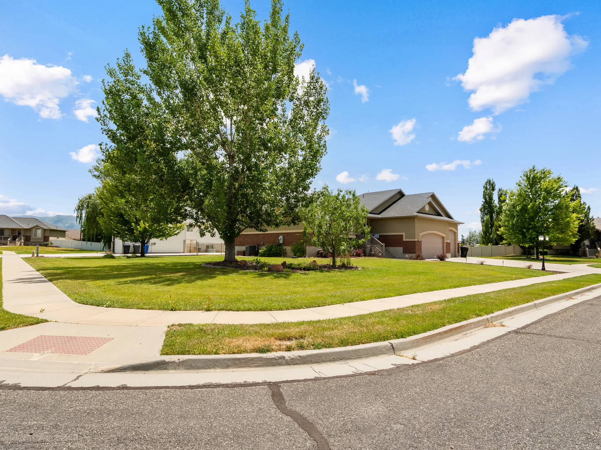 Spacious corner lot featuring a manicured lawn, established shade trees, paved sidewalks, and a concrete driveway leading to an attached garage