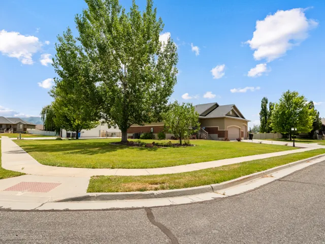 Spacious corner lot featuring a manicured lawn, established shade trees, paved sidewalks, and a concrete driveway leading to an attached garage