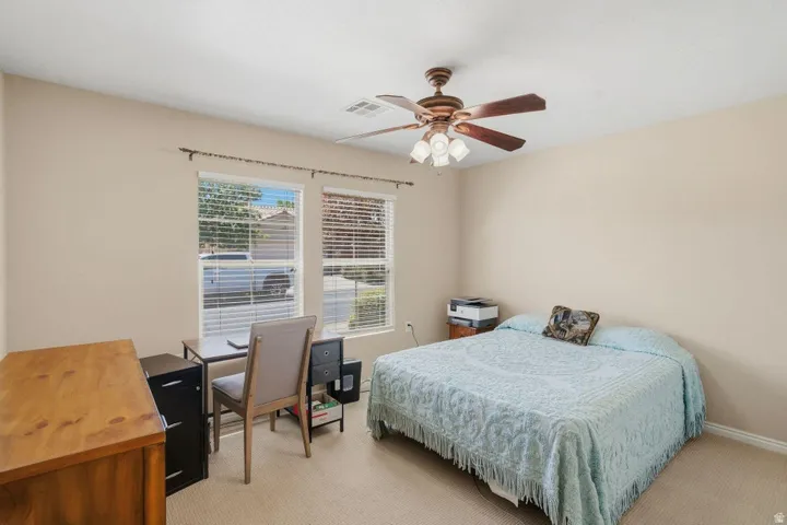 Neutral tone wall paint, textured carpet flooring, two windows with horizontal blinds, and a ceiling fan with integrated lighting