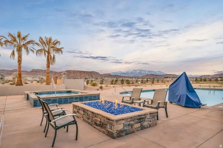 Spacious outdoor patio featuring a stone-clad fire pit with blue fire glass, two hot tubs with blue tile accents and water features, a swimming pool, and desert landscaping