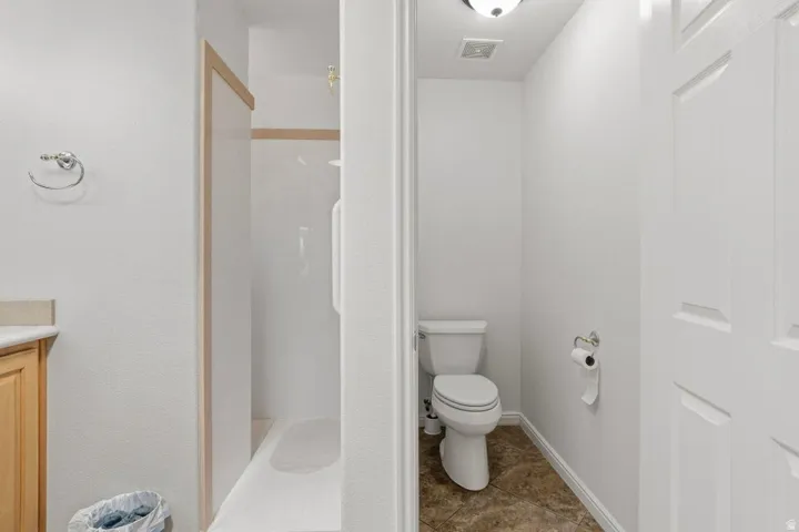 Bathroom featuring a walk-in shower with a white surround, a separate water closet with a commode, a light wood-finish vanity with a light-toned countertop, and a chrome towel ring