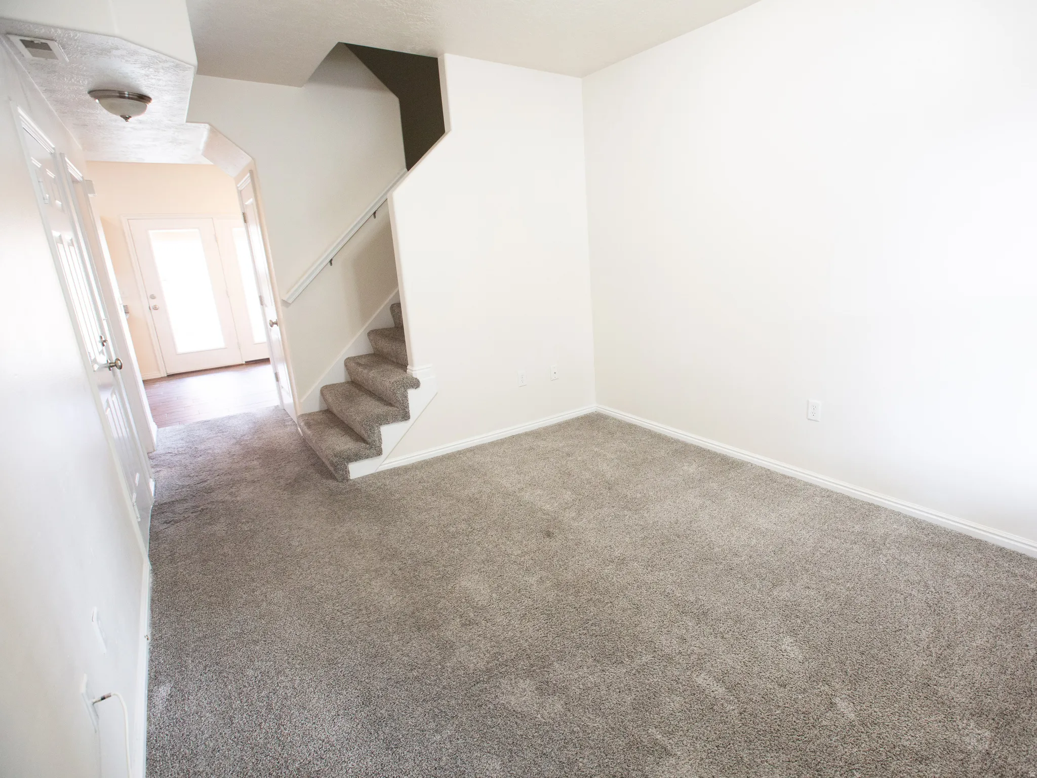 Neutral gray carpeting throughout, featuring white baseboards, a staircase with white railing, and a hallway leading to exterior glass-paneled doors with wood-finish flooring