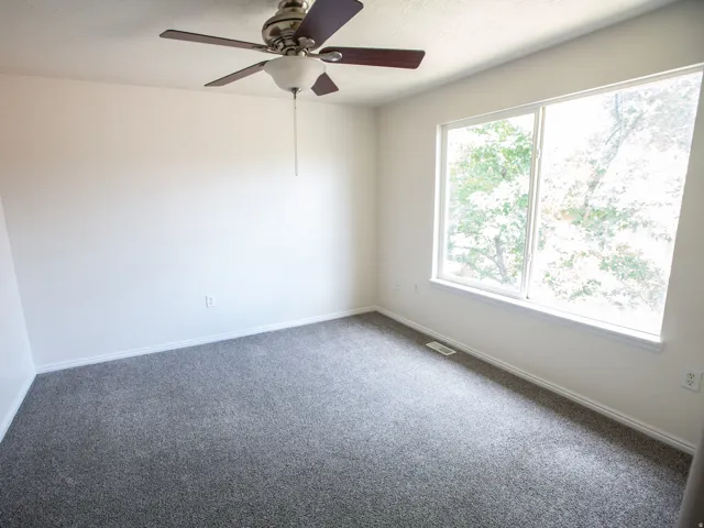 Bright room featuring a large window, gray carpeting, a ceiling fan with integrated lighting, white walls, and white baseboards