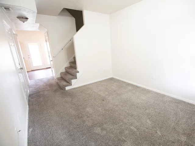 Neutral gray carpeting throughout, featuring white baseboards, a staircase with white railing, and a hallway leading to exterior glass-paneled doors with wood-finish flooring