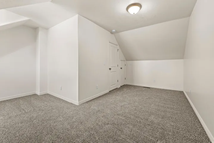 Spacious room featuring sloped ceilings, plush gray carpeting, white baseboards, and two white doors with dark hardware