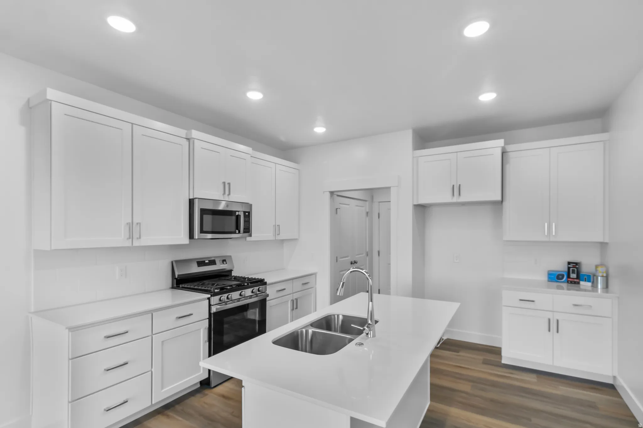 Bright kitchen featuring white shaker cabinetry, stainless steel appliances, a center island with a double basin sink, recessed lighting, and wood-finish flooring