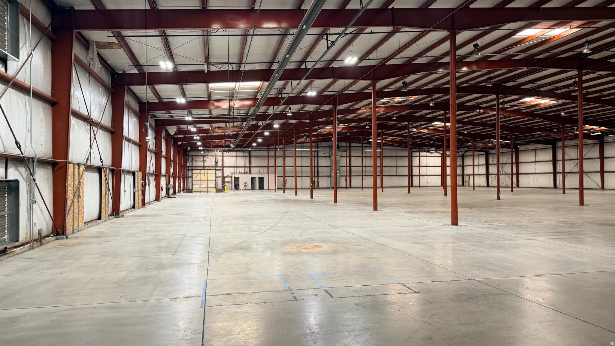 Expansive industrial warehouse featuring a vast concrete floor, high ceilings with exposed red structural beams, and numerous overhead lighting fixtures