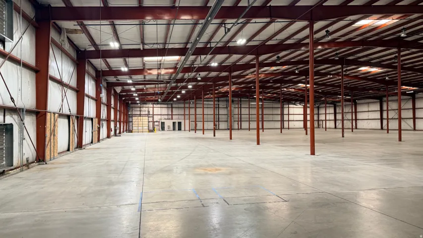 Expansive industrial warehouse featuring a vast concrete floor, high ceilings with exposed red structural beams, and numerous overhead lighting fixtures