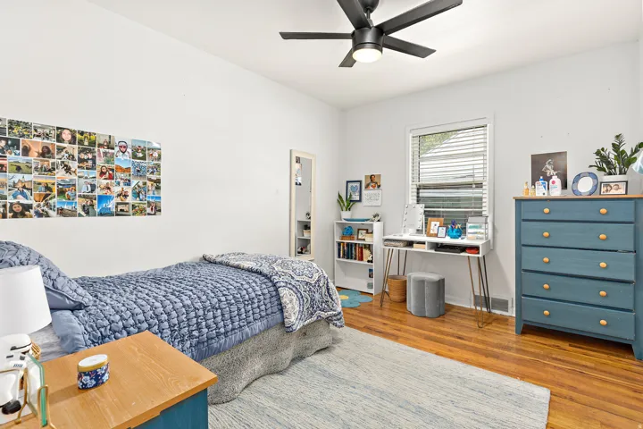 Hardwood flooring throughout this bright room featuring a ceiling fan with integrated lighting, a window with horizontal blinds, and a built-in bookshelf