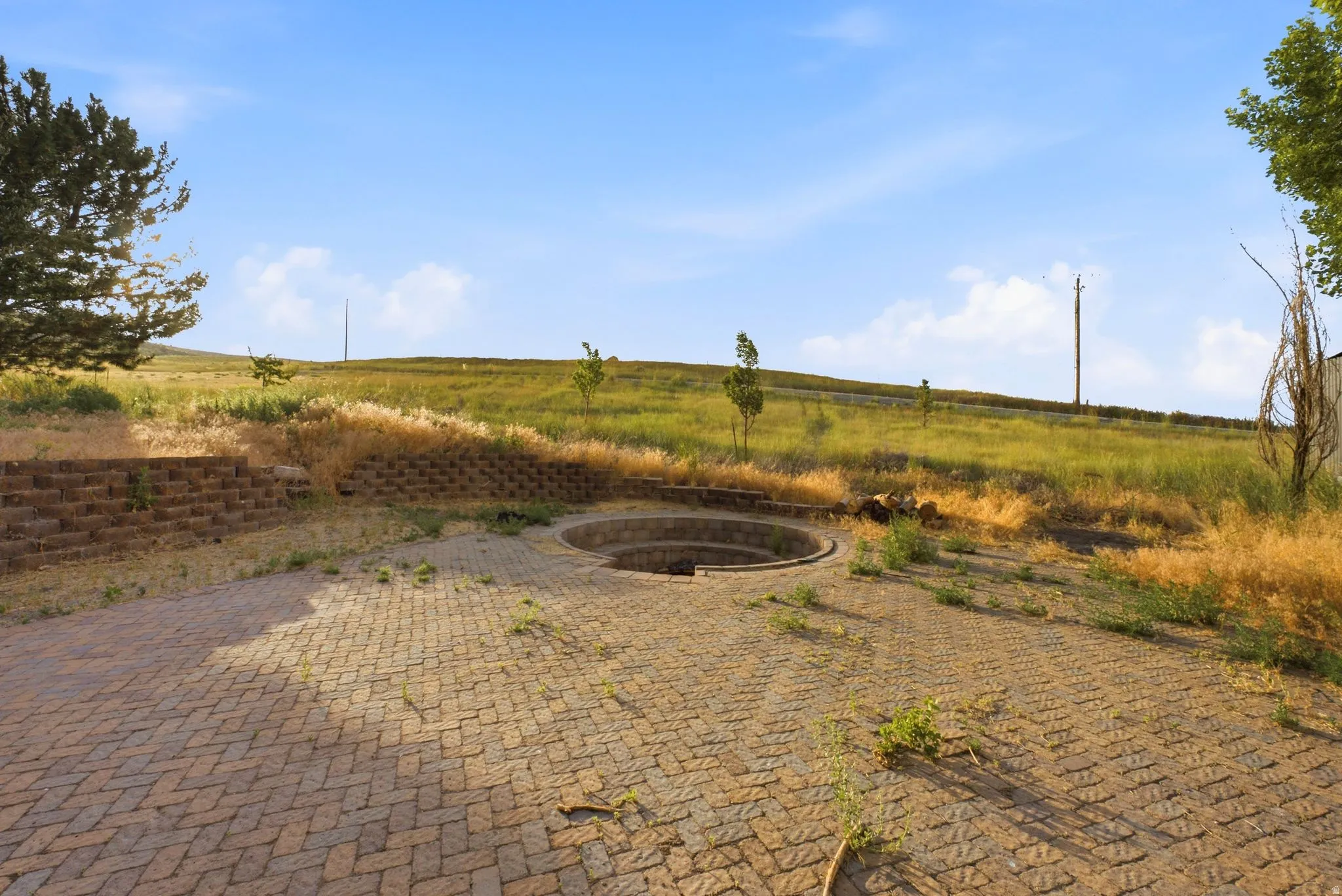 Paved outdoor area featuring a circular recessed fire pit, block retaining walls, and a gently sloping landscape