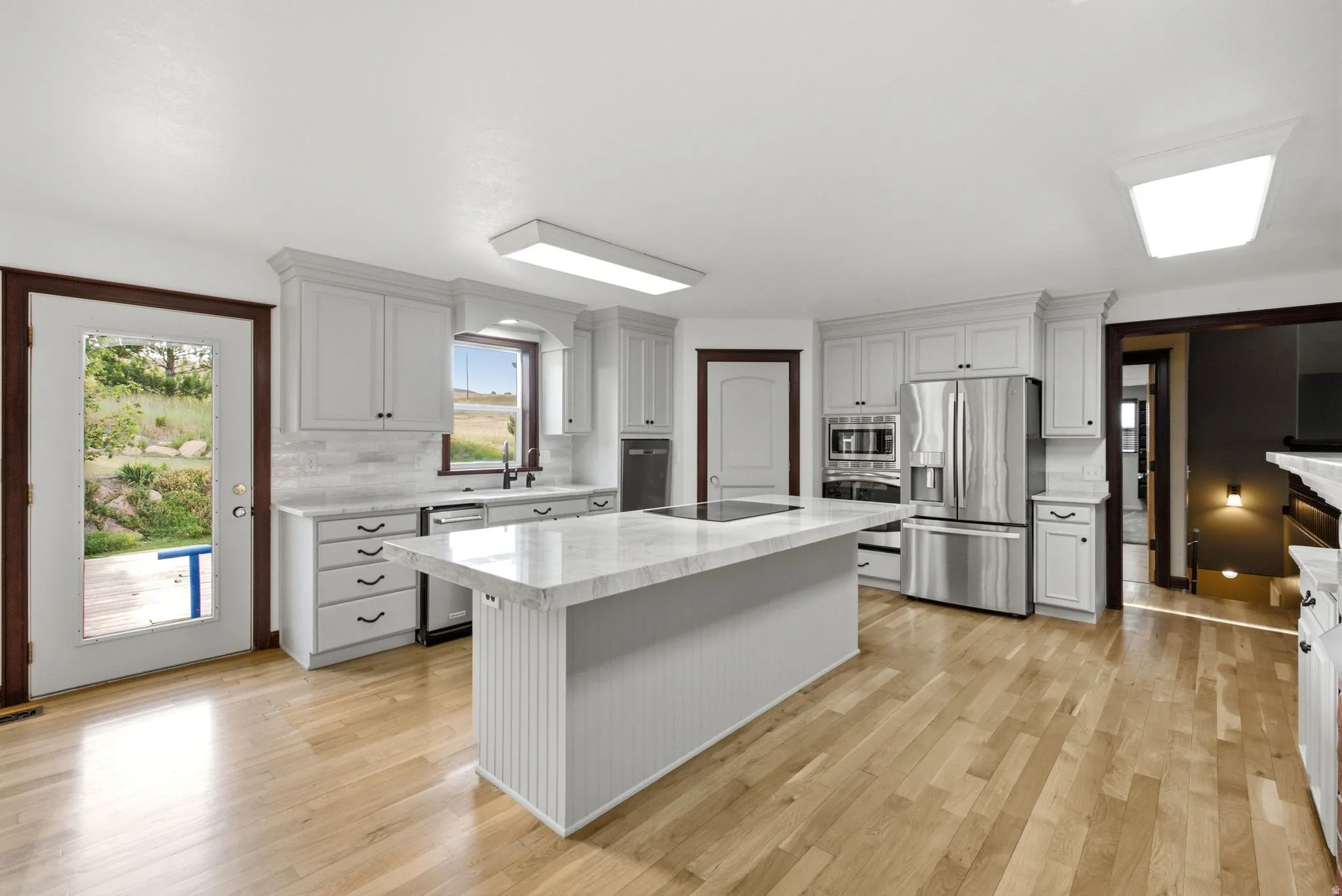 Spacious kitchen featuring an expansive center island with an integrated cooktop, light-toned wood-finish flooring, white shaker-style cabinetry, and stainless steel appliances