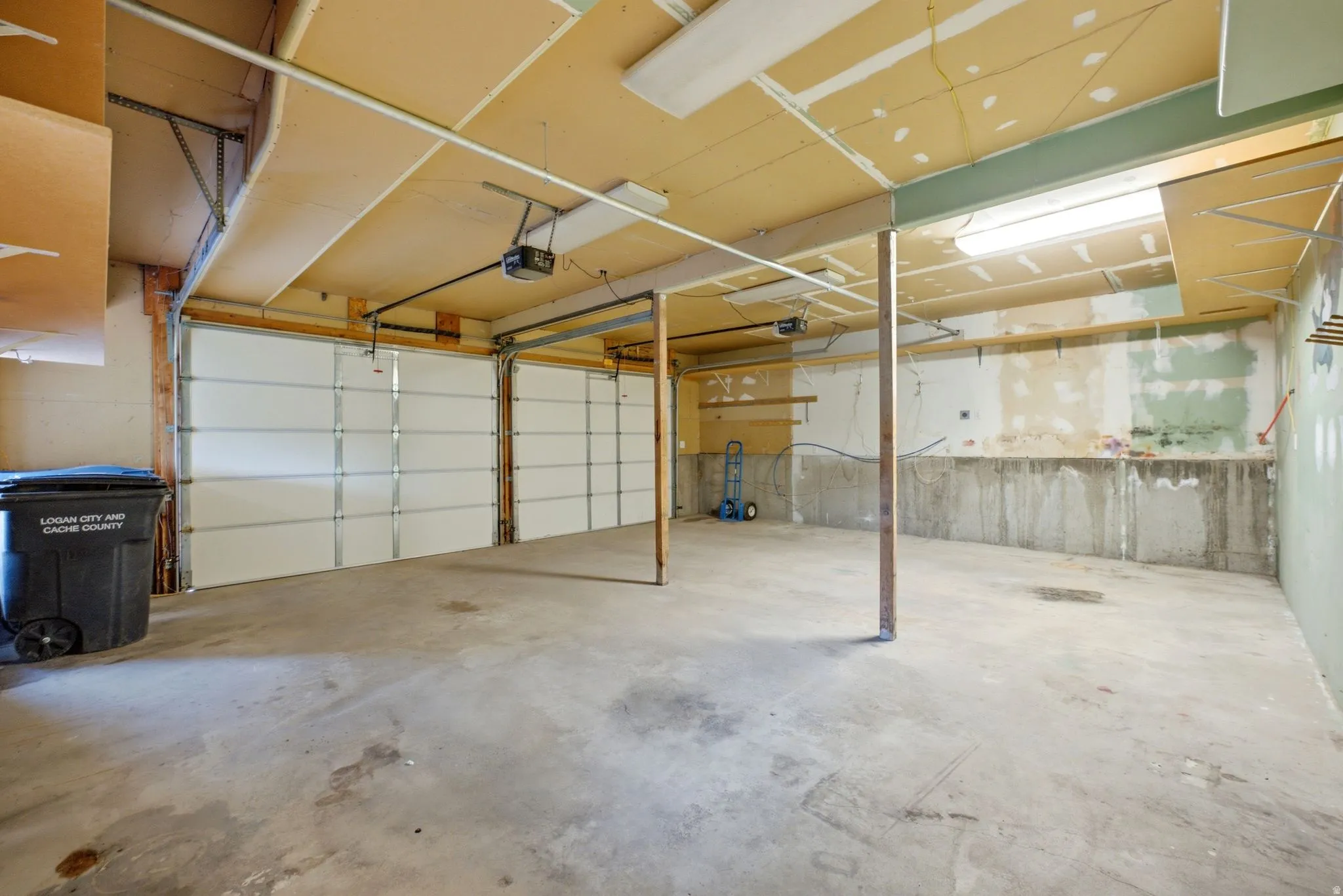 Main home basement. Spacious garage with concrete flooring and exposed ceiling joists, featuring two white panel garage doors, track lighting, and a concrete foundation wall