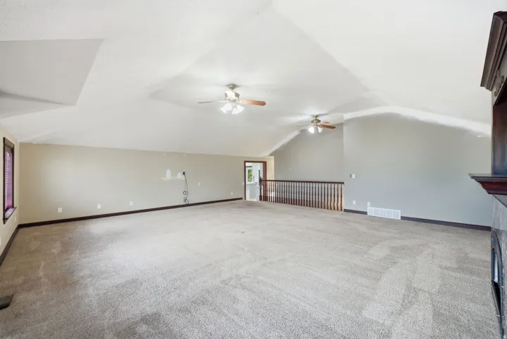 Spacious carpeted room featuring a vaulted ceiling with two ceiling fans, a dark wood window frame, and an open railing overlooking a lower level