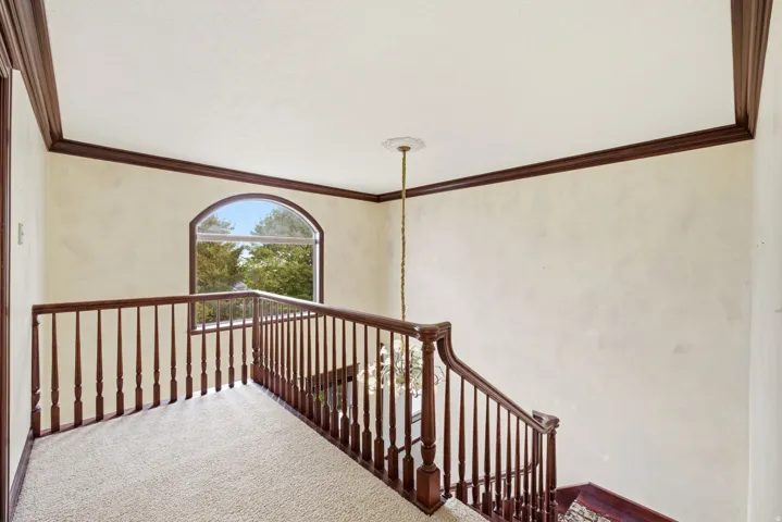 Carpeted staircase and landing featuring dark wood railings and balusters, an arched window, and decorative crown molding