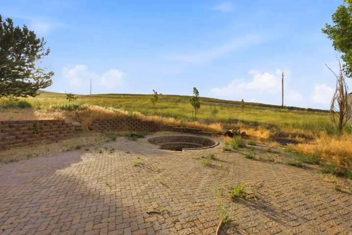 Paved outdoor area featuring a circular recessed fire pit, block retaining walls, and a gently sloping landscape