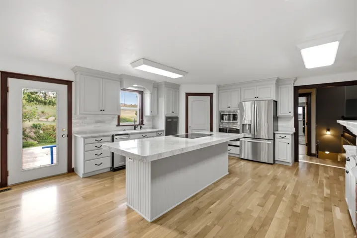 Spacious kitchen featuring an expansive center island with an integrated cooktop, light-toned wood-finish flooring, white shaker-style cabinetry, and stainless steel appliances