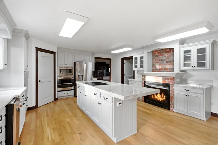 Kitchen featuring light wood-finish flooring, a central island with a cooktop, white perimeter cabinetry, stainless steel appliances, and a brick-accented fireplace