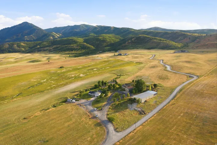 Expansive rural property featuring a winding paved road, multiple residences, a large agricultural building, and surrounding mountainous terrain