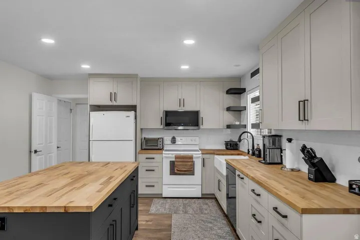 Modern kitchen featuring a central island with wood-finish countertop, light-colored cabinetry with matte black hardware, recessed lighting, and a farmhouse sink