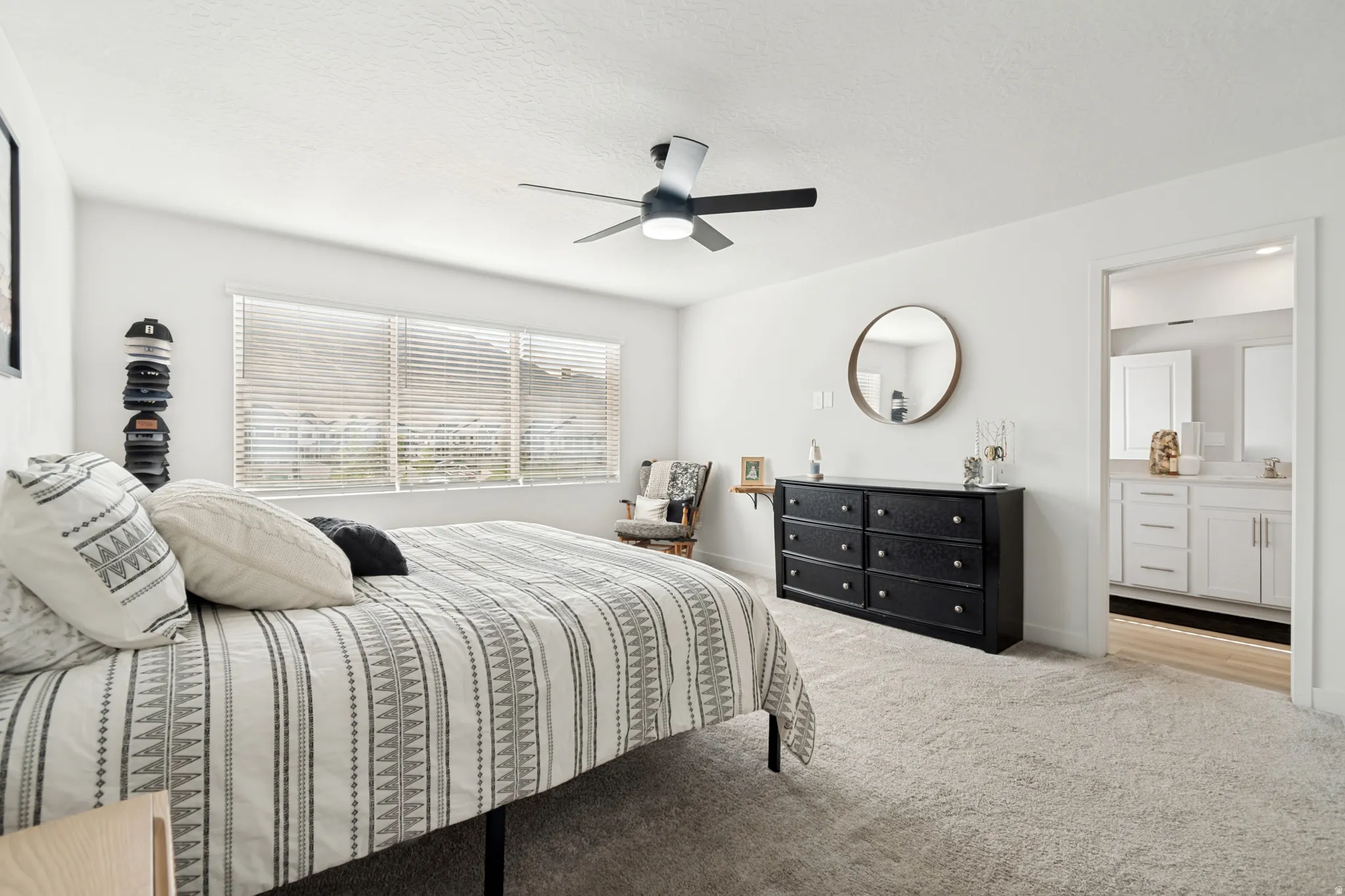 Carpeted bedroom featuring a ceiling fan with integrated lighting, a large window with horizontal blinds, and a doorway opening to a bathroom with a dual vanity