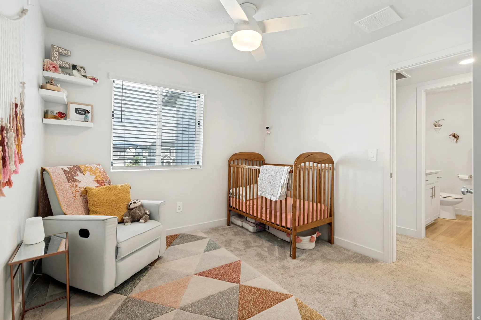 Carpeted room featuring a ceiling fan with integrated lighting, a window with horizontal blinds, built-in wall shelving, and an adjacent bathroom with wood-finish flooring
